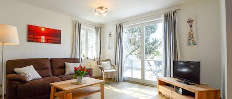 A living room with a brown sofa, a wooden coffee table, a TV on a wooden stand, and a large window with curtains.
