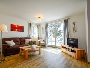 A living room with a brown sofa, a wooden coffee table, a TV on a wooden stand, and a large window with curtains.