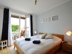 Un dormitorio con una cama grande, una mesita de noche de madera, un escritorio y una silla. Hay un ventanal corredero que da a un área exterior.