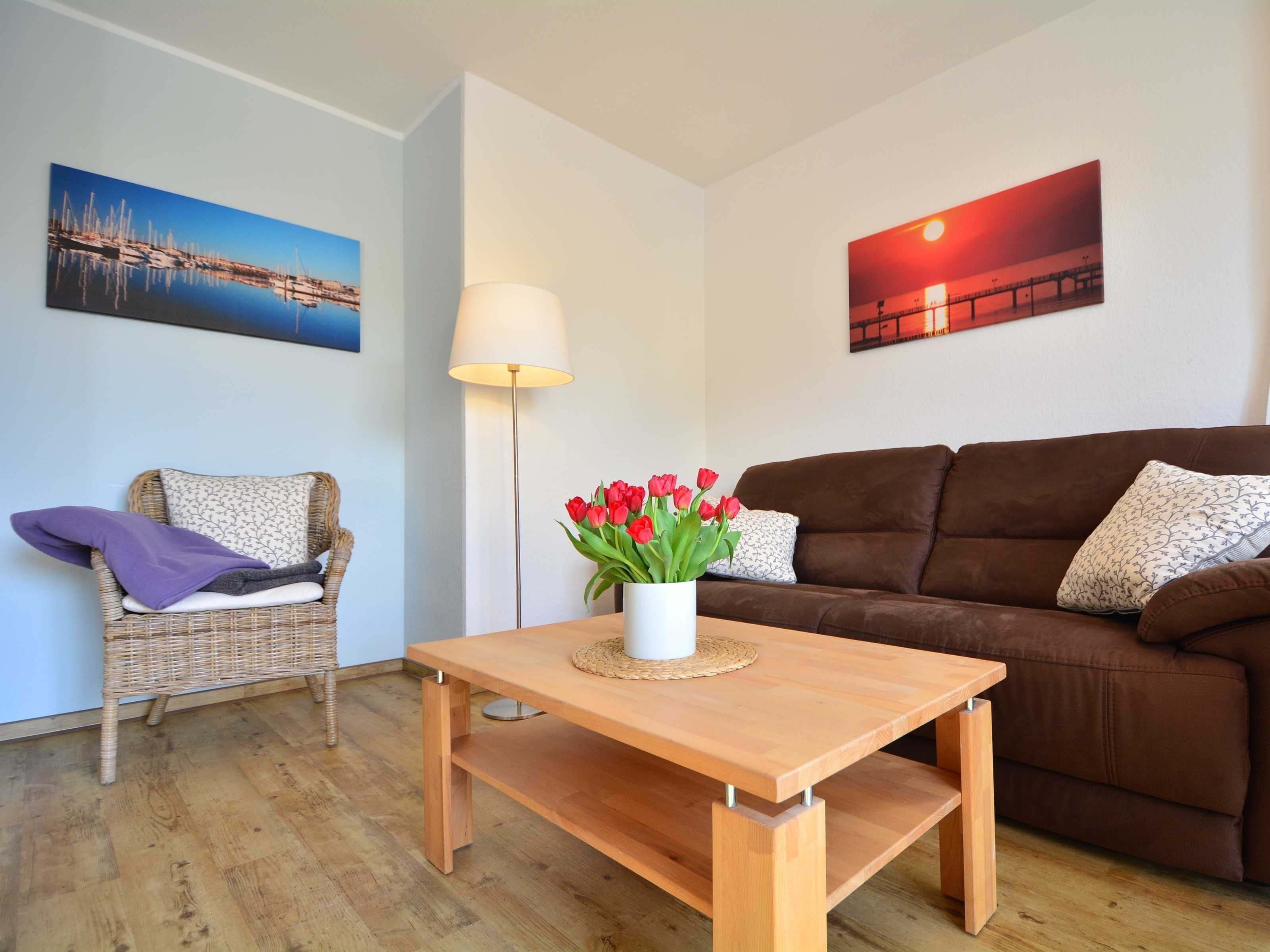 A living room with a wooden table, a brown sofa, a wicker chair, and two wall-mounted paintings.