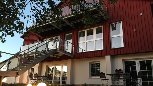 A two-story building with a red exterior, a balcony with metal railings, and a patio area with outdoor seating and lighting. - Apartment 1 - Usedom Landhaus Morgenitz (Mellenthin OT Morgenitz)