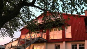A red two-story house with a staircase leading to the upper floor, surrounded by greenery and a clear sky. - Apartment 1 - Usedom Landhaus Morgenitz (Mellenthin OT Morgenitz)
