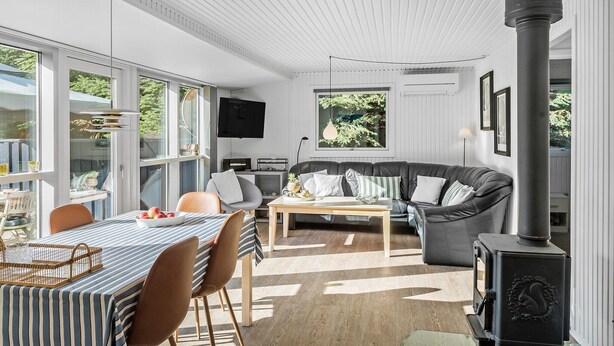 A modern living room with a large window, a sofa, a dining table, and a wood-burning stove. - Pet friendly home in Væggerløse with sauna (Væggerløse)