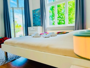 A bed with a white mattress and pillows, a window with a view of greenery, and a lamp on the bedside table.