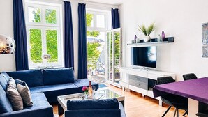 A modern living room with a blue sofa, a glass coffee table, a dining table with chairs, a TV, and a chandelier.