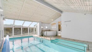 A modern indoor swimming pool with clear glass walls, a jacuzzi, and a view of the outdoors.