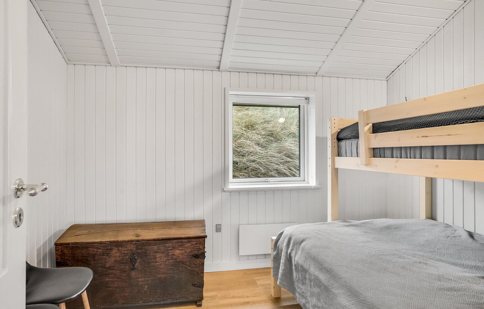 A bedroom with a bunk bed, a wooden chest, a chair, and a window with a view of trees.