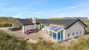 A modern beach house with a large glass-enclosed patio, a dining area with outdoor seating, and a separate lounge area with purple cushions.