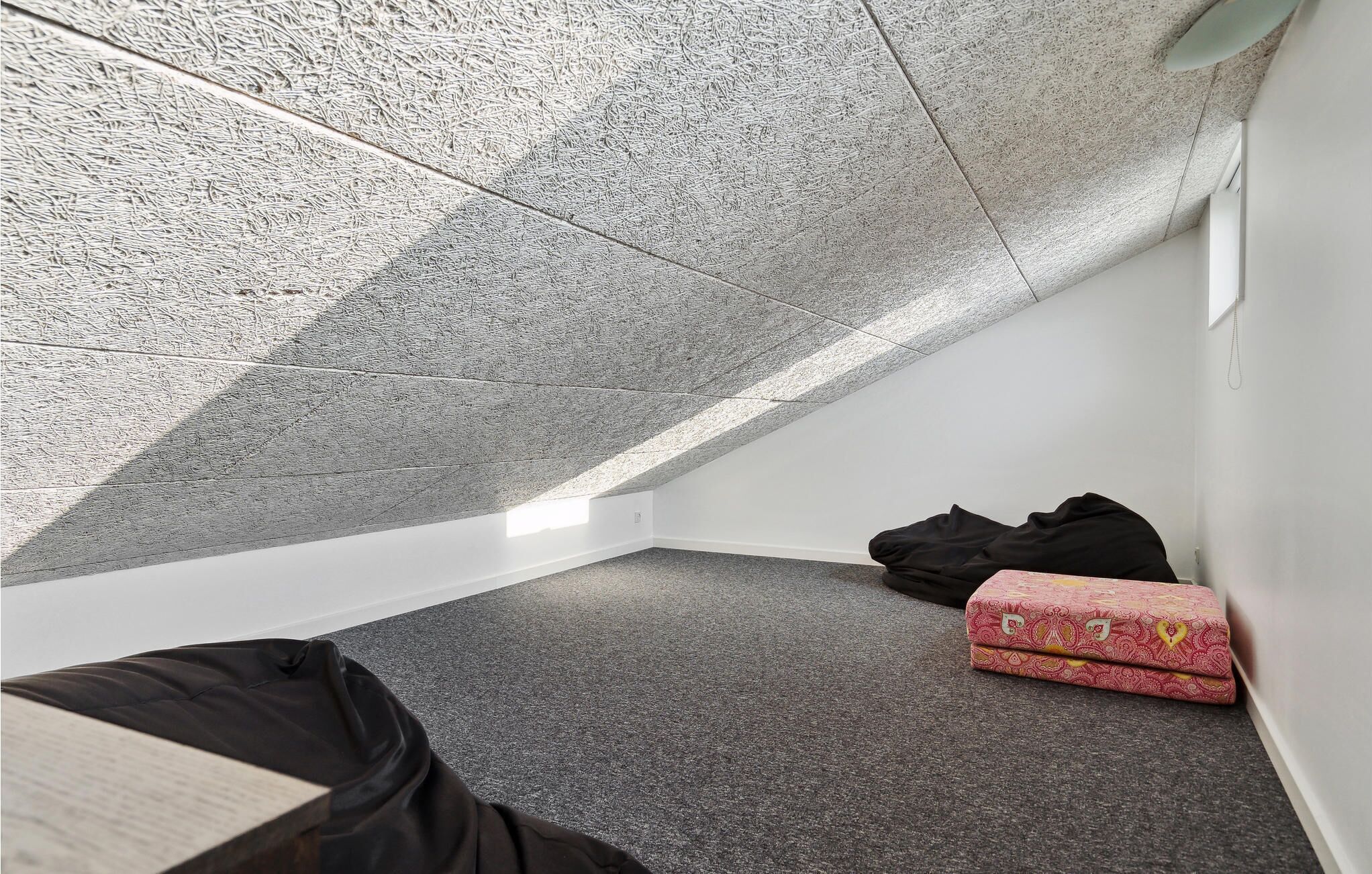 A small, dimly lit attic room with a sloped ceiling, a single window, and a carpeted floor.