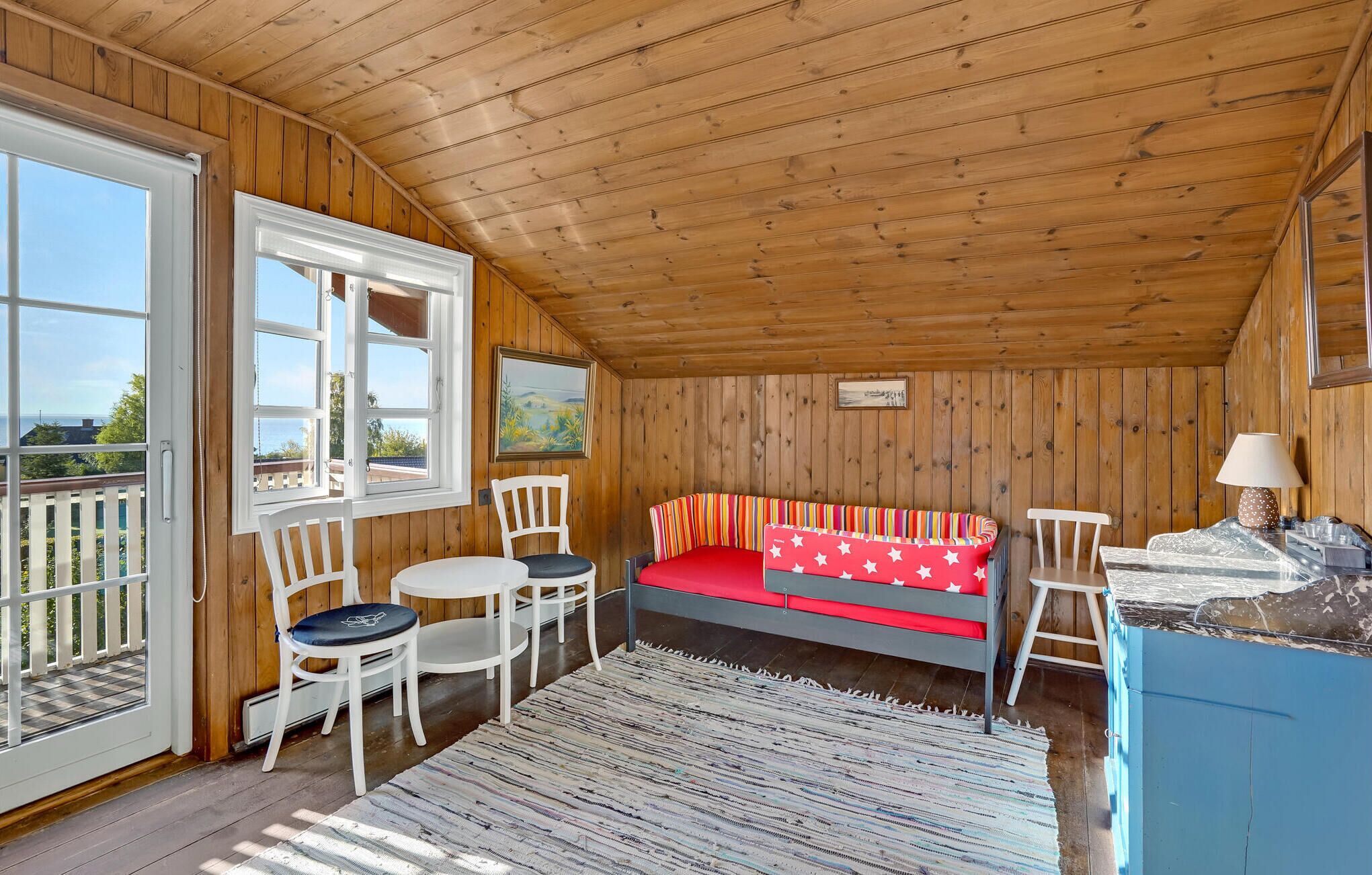 A cozy cabin with a wooden interior, a bed with a colorful striped blanket, a small dining table with chairs, and a view through the open door.
