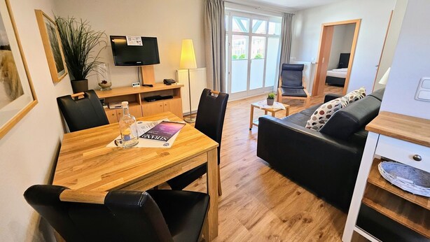 Een moderne woonkamer met een houten eettafel, zwarte lederen stoelen, een zwarte bank met gekleurde kussens, een televisie en een lamp.