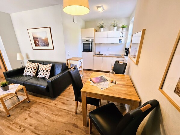 A modern living room with a black leather sofa, a wooden dining table, and a kitchen area with white cabinets and a built-in oven.