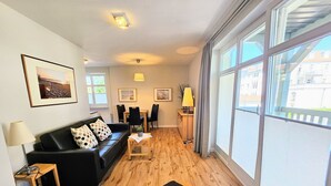 Een moderne woonkamer met een zwarte leren bank, een houten salontafel en een eethoek met stoelen. Grote ramen bieden uitzicht op het exterieur.