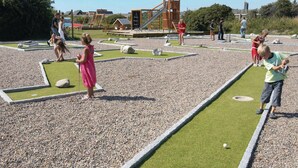 A children's play area with a mini-golf setup, a wooden play structure, and a small building in the background. - 2 bedroom beach front home in Thyborøn (Thyborøn)