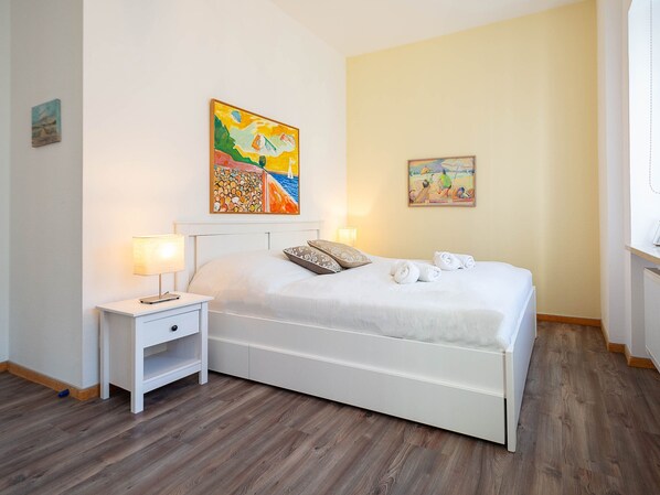 Un dormitorio con una cama blanca, dos mesitas de noche con lámparas y dos cuadros en la pared.