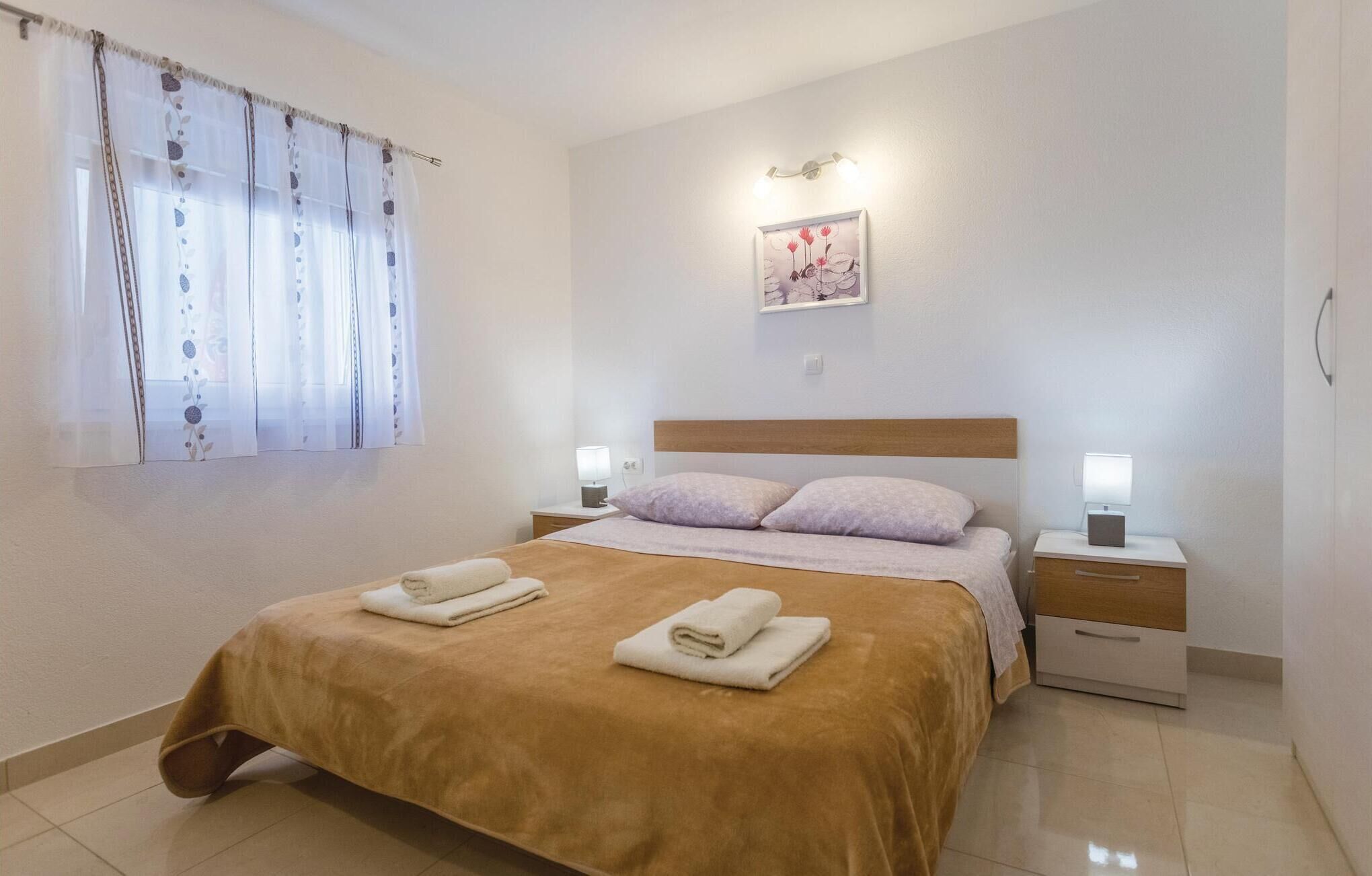 A hotel room with a bed, two bedside tables, a nightstand, a window with curtains, and a wall-mounted light fixture.