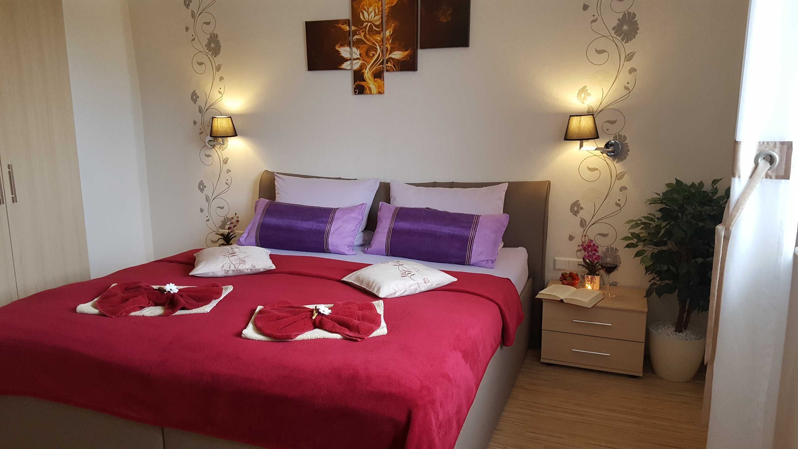 Ein ordentlich bezogenes Bett mit roter Tagesdecke, zwei violetten und einem weißen Kissen. An beiden Seiten des Bettes befinden sich zwei Wandlampen.