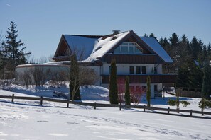 A two-story house with a sloped roof, surrounded by snow-covered ground and trees.