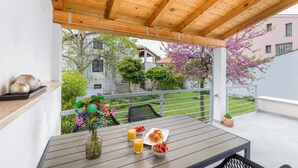 Een overdekt terras met een ontbijttafel, compleet met croissants en fruit, en een bloevas.