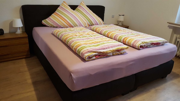 A bed with a striped blanket and pillows, a bedside table with a lamp, and a wooden floor.