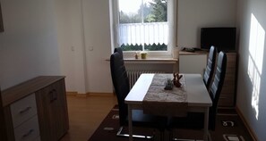 Un coin repas comprenant une table, des chaises, un radiateur, une fenêtre avec des rideaux, un téléviseur et un meuble.