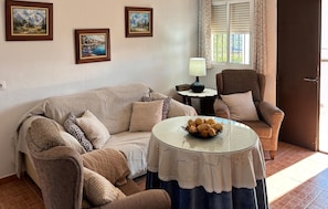 Un salon chaleureux avec un canapé à motifs, une table basse ronde sur laquelle se trouve un bol de collations, et un fauteuil avec un coussin. Trois tableaux encadrés ornent le mur, et une lampe est posée sur une petite table.