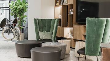 Ein modernes Wohnzimmer mit grün gepolsterten Sesseln, einer Couch und Poufs. Ein Bücherregal mit verschiedenen Gegenständen und ein Fernseher sind ebenfalls zu sehen. Im Hintergrund ist ein Fahrrad erkennbar.