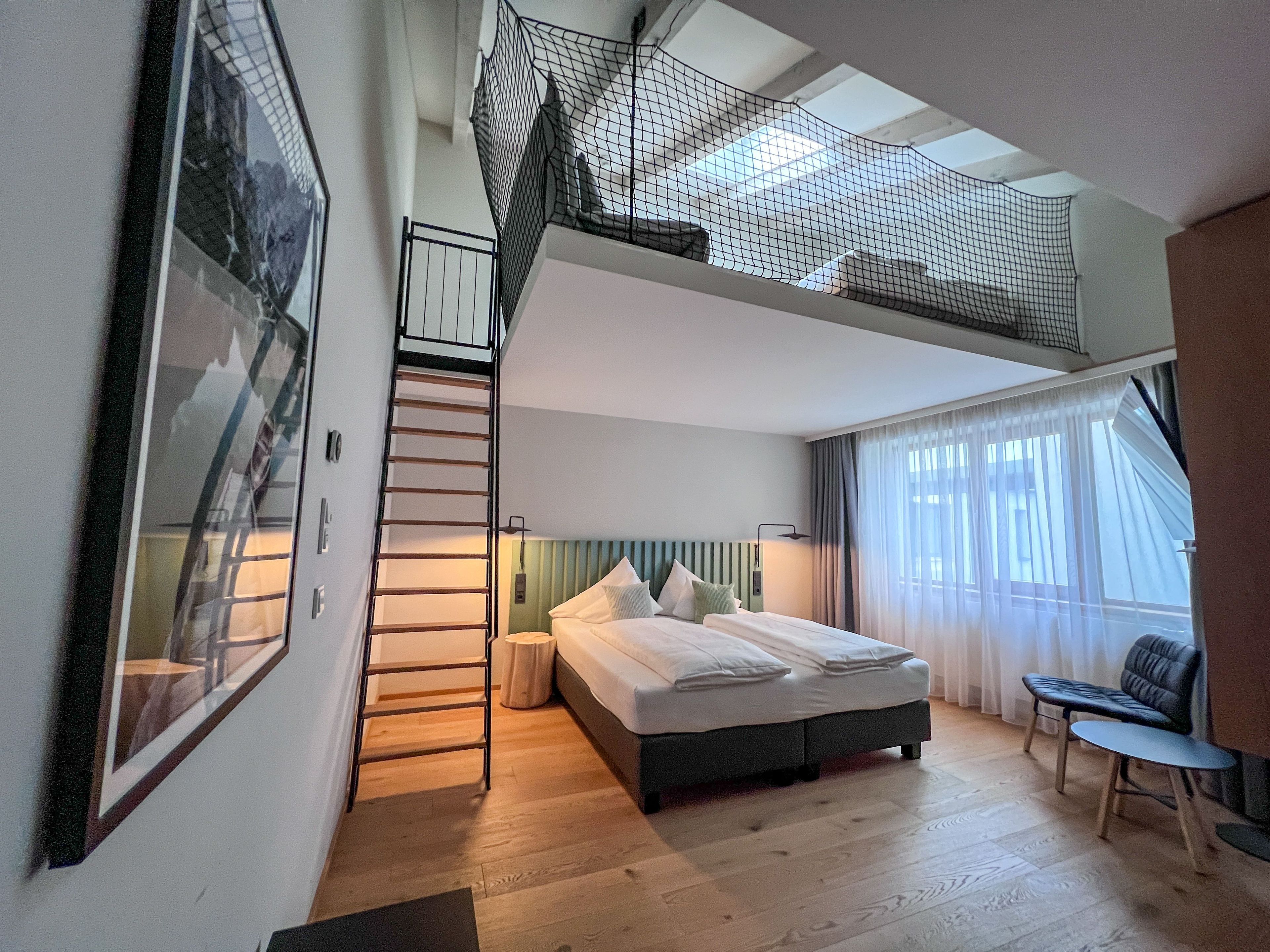 Une chambre à coucher comprenant un lit, un bureau, une chaise, une fenêtre avec des rideaux, un escalier et un balcon muni d’un filet de protection.