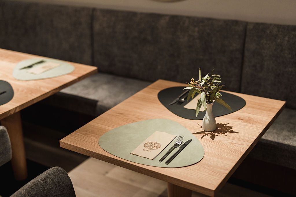 Ein Restaurant mit gedeckten Tischen, jeweils mit Untersetzer, Besteck und einer kleinen Vase mit Blumen.