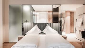A modern hotel room with a large bed, a mirror, a wooden wardrobe, and a bathroom visible through a glass partition.