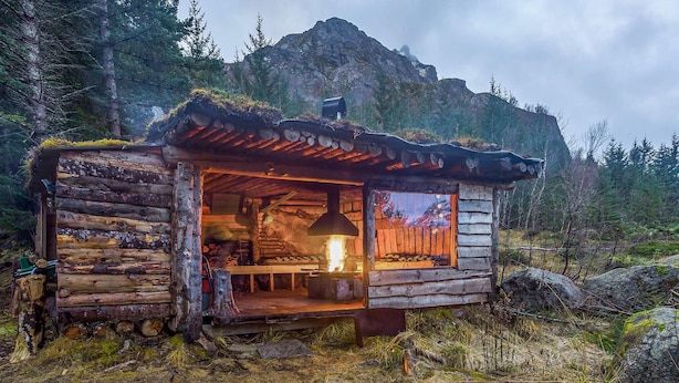 Una cabaña rústica con fachada de madera, chimenea central y vistas al bosque circundante.