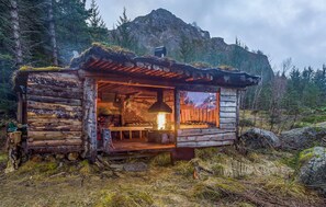 Una cabaña rústica con fachada de madera, chimenea central y vistas al bosque circundante.