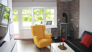 Un salón con un sillón amarillo, un sofá negro con un cojín rojo, una chimenea y un amplio ventanal con vistas a la vegetación.