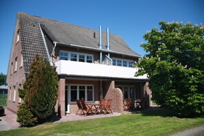 Une maison à deux étages avec une façade en briques, un patio couvert avec des meubles d’extérieur et un grand arbre dans le jardin.