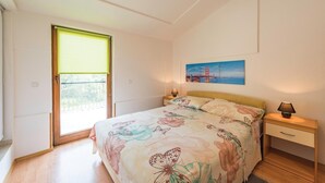 Un dormitorio con cama, mesita de noche, lámpara, un cuadro de un puente y una ventana con vistas a la vegetación.