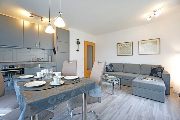 Une cuisine moderne avec des meubles gris, une table à manger dressée avec des assiettes et des verres, et un canapé gris dans l’espace salon.