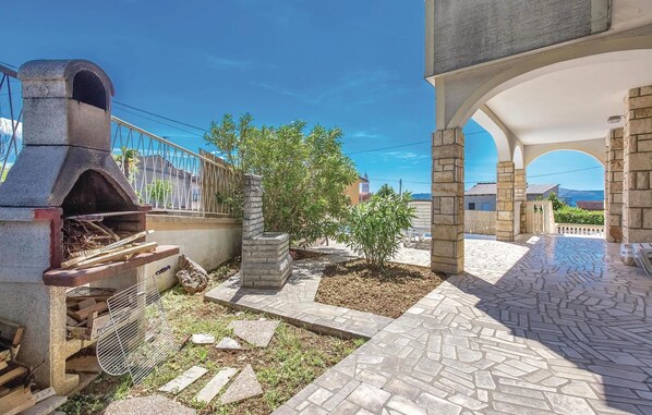 Un patio con una parrilla de piedra, un árbol pequeño y un camino pavimentado que lleva a un edificio con una entrada arqueada.