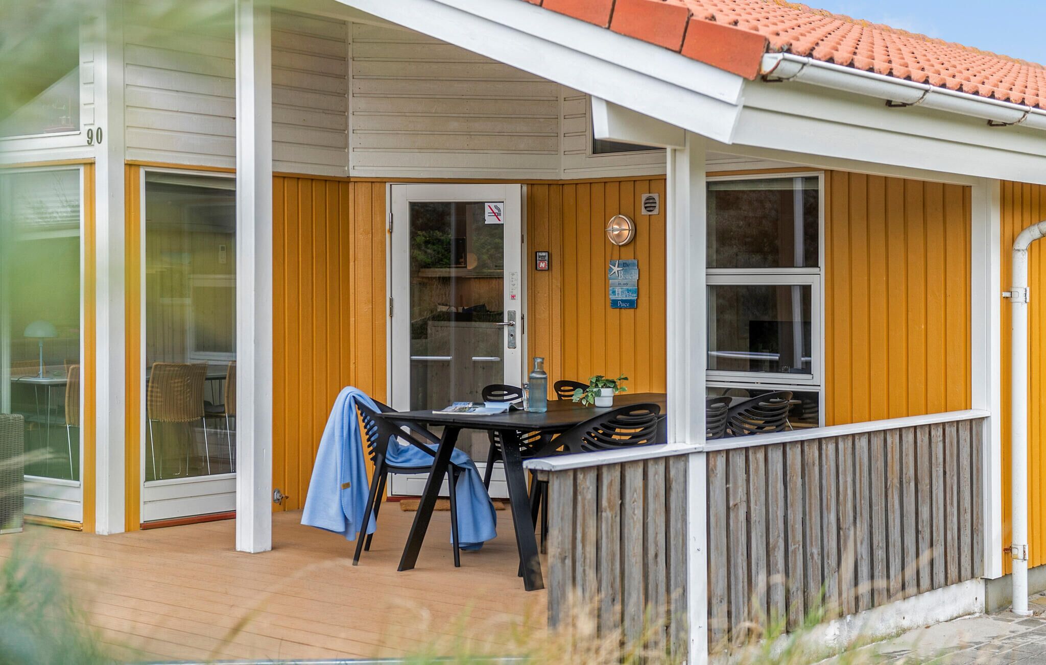 Ett takförsett utomhusområde med ett bord och stolar, ett trästaket och en byggnad med gul fasadbeklädnad och ett tak täckt med takpannor.