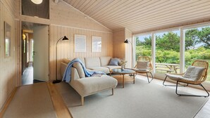 A cozy room with a sofa, armchairs, and a dining table, featuring wooden walls and large windows.