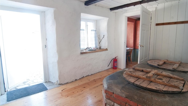 A room with wooden flooring, a brick fireplace, a window with a plant, and a door leading to another room.