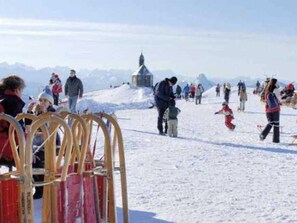 Un paisaje nevado con gente caminando y parada, un pequeño edificio y un telesilla.