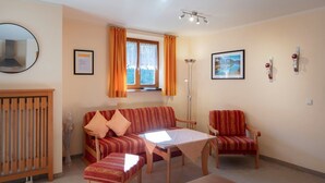 A living room with a sofa, two armchairs, a coffee table, a wooden bench, a mirror, a window with curtains, a floor lamp, and two framed pictures on the wall. - Apartment 7 - Apartments Lohbinder (Fam. Holzer) (Rottach-Egern)