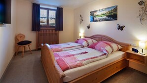 A bedroom with a wooden bed, a chair, a nightstand with a lamp, a window with curtains, and wall decorations. - Apartment 7 - Apartments Lohbinder (Fam. Holzer) (Rottach-Egern)