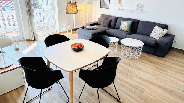 A modern living room with a grey sofa, a white dining table, black chairs, and a bowl of fruit. There is a window with curtains, a lamp, and a framed picture on the wall. - Barbarossa Whg B-12. - B-12 (Kühlungsborn)