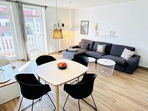 A modern living room with a grey sofa, a white dining table, black chairs, and a bowl of fruit. There is a window with curtains, a lamp, and a framed picture on the wall. - Barbarossa Whg B-12. - B-12 (Kühlungsborn)