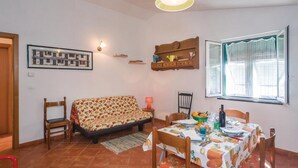 A cozy living room with a sofa, chairs, a dining table set for two, a framed picture on the wall, and a window with blinds. - Lovely home in Moneglia GE with WiFi (Moneglia GE)