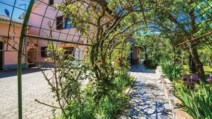 A garden pathway leading to a building with a pink facade, surrounded by greenery and a clear blue sky. - Lovely apartment in Matulji (Matulji)