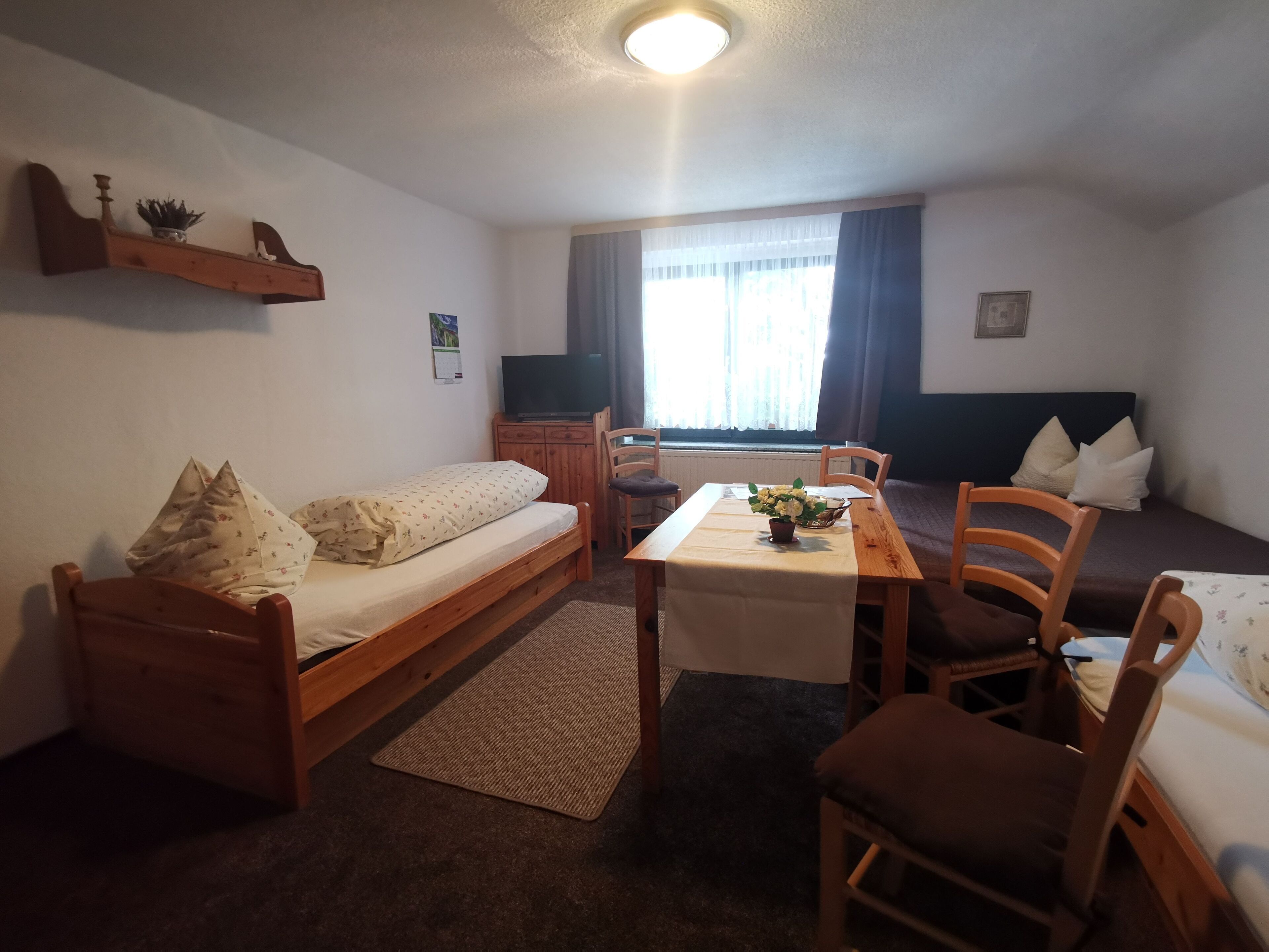 A small, cozy room with a bed, a desk, and a chair. There is a window with curtains, a radiator, and a shelf with decorative items.