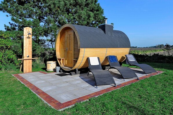 A wooden barrel-shaped structure with a door, a small wooden table, and two lounge chairs on a paved patio.