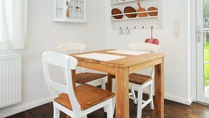 Un comedor con mesa de madera y sillas blancas, una lámpara colgante blanca, un mueble con vajilla y una ventana con cortinas.